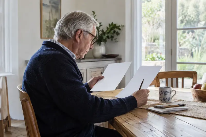 Lector mayor ilustrativo revisando hojas de comparación sencillas en una mesa de comedor.