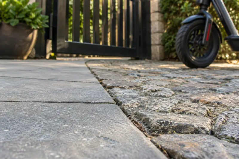 Illustrative detail of smooth paving meeting textured stone beside a gate and stationary wheel.